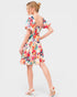 A woman modeling a short puff-sleeve dress from the back, featuring a vibrant multicolor floral print on a cream background, paired with white heeled sandals against a light gray studio background.