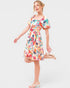 A woman modeling a short puff-sleeve dress with a colorful floral print on a light background, paired with white heeled sandals, against a light gray studio backdrop.