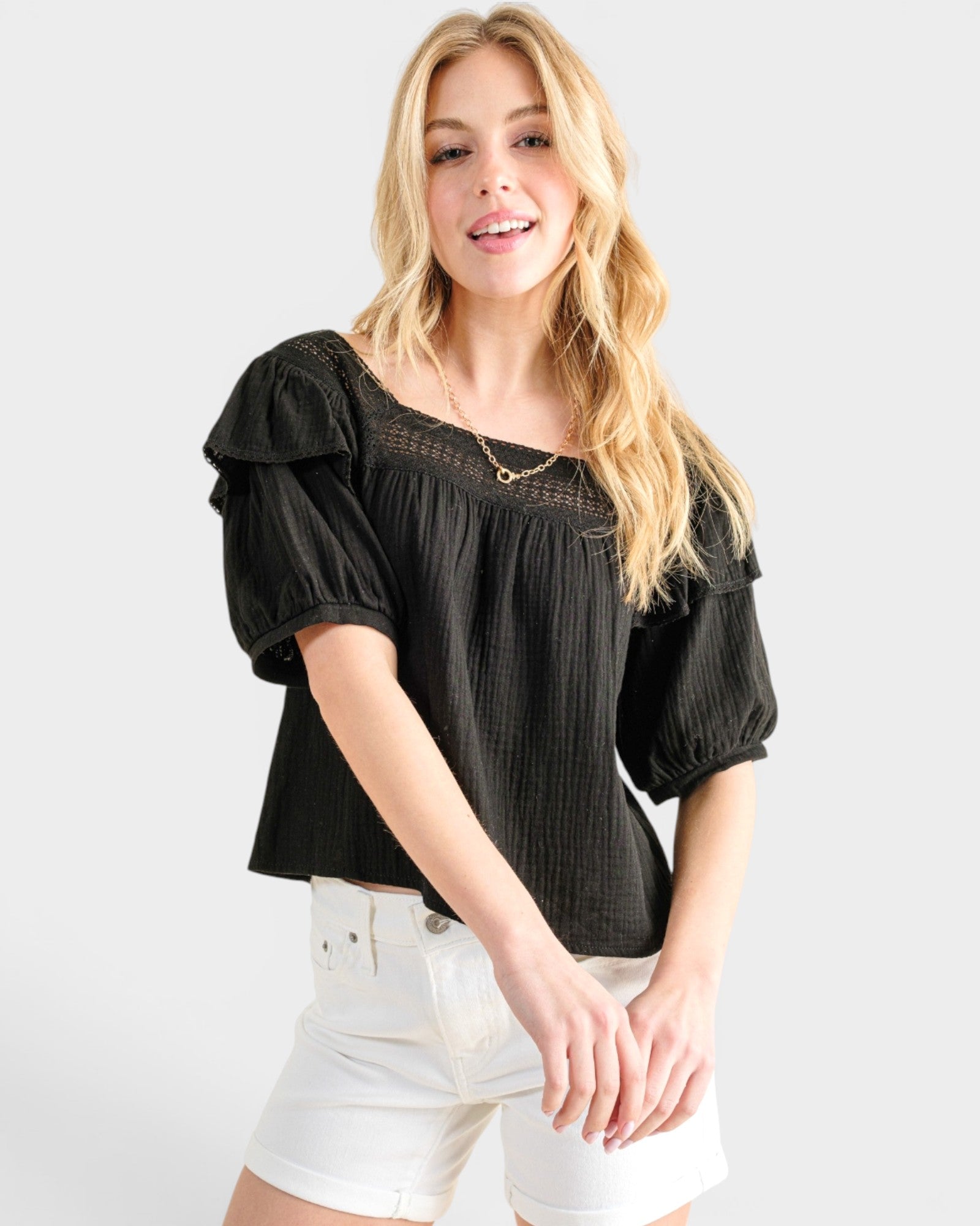 Woman wearing a black blouse with ruffled sleeves and white shorts on a light gray background