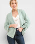 Woman wearing a light green blazer over a white top and blue jeans on a plain background