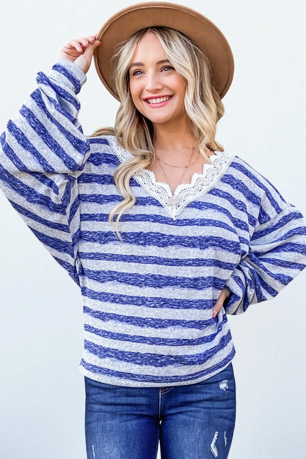 Woman wearing a blue and white striped sweater with lace details, blue jeans, and a brown hat on a light background.