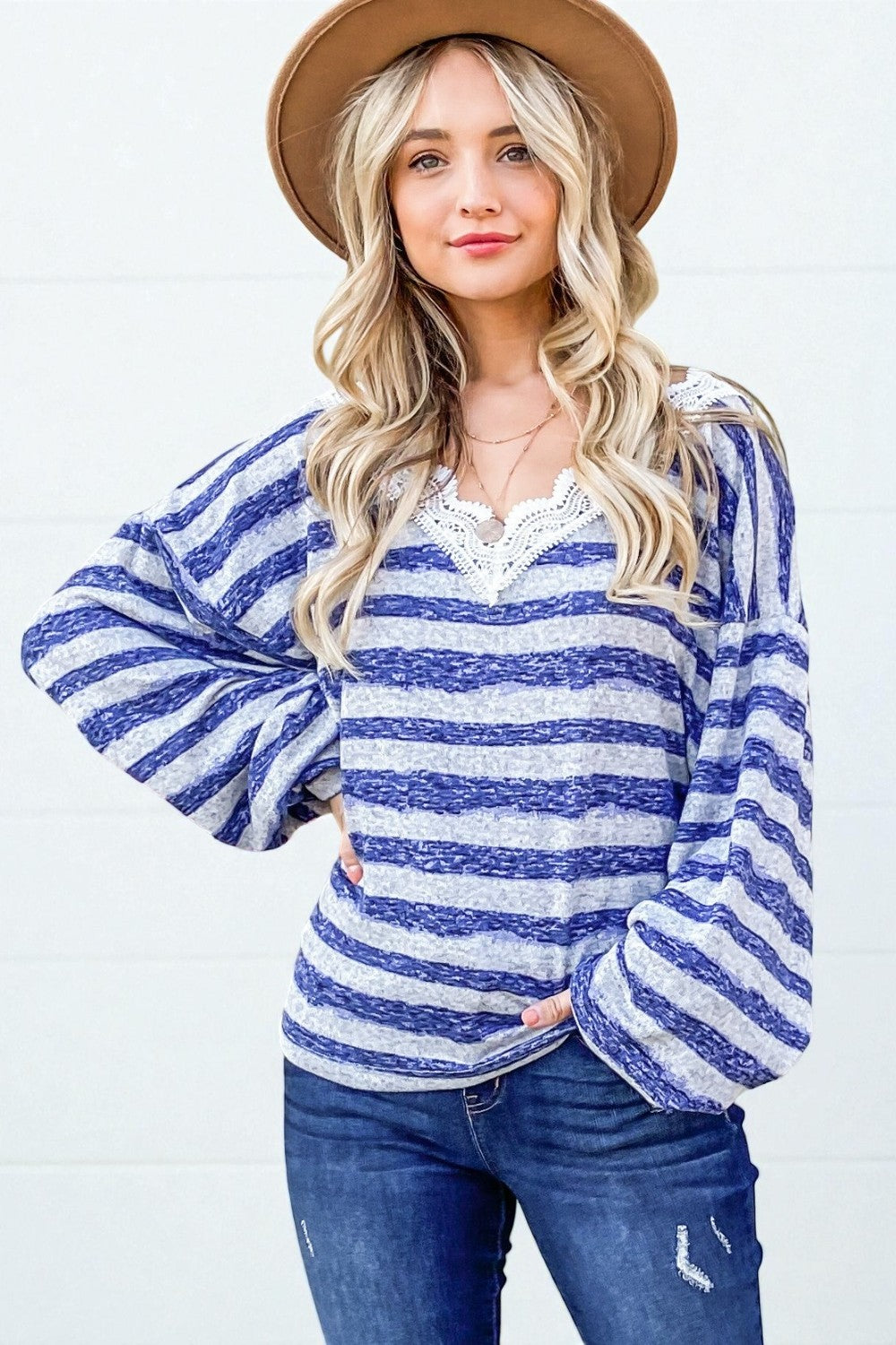 Woman wearing a blue and white striped long-sleeve top with a lace neckline, jeans, and a brown hat on a white background.