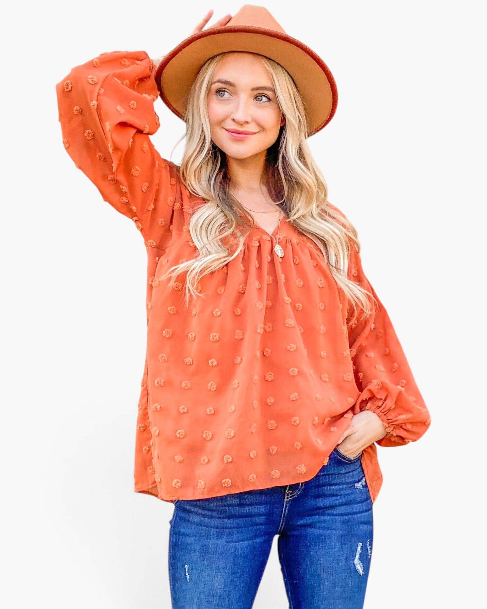 A woman modeling a long-sleeve terracotta orange blouse with a textured raised-dot pattern, paired with blue jeans and a brown wide-brim hat, against a plain white background.