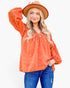 A woman modeling a long-sleeve terracotta orange blouse with a textured raised-dot pattern, paired with blue jeans and a brown wide-brim hat, against a plain white background.