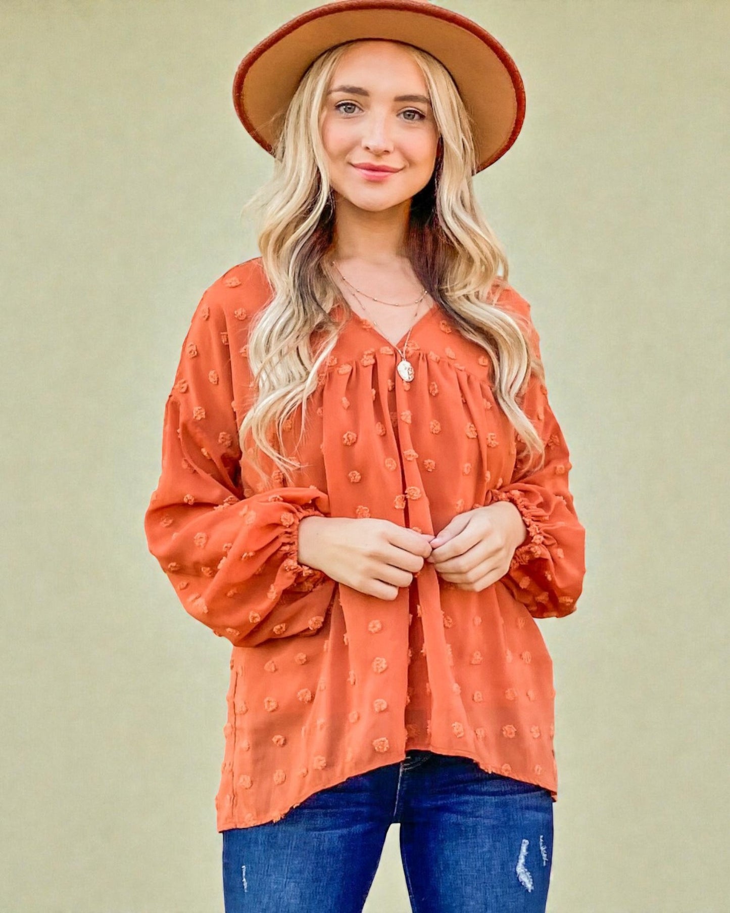 Woman wearing an orange blouse and blue jeans with a beige hat against a beige background