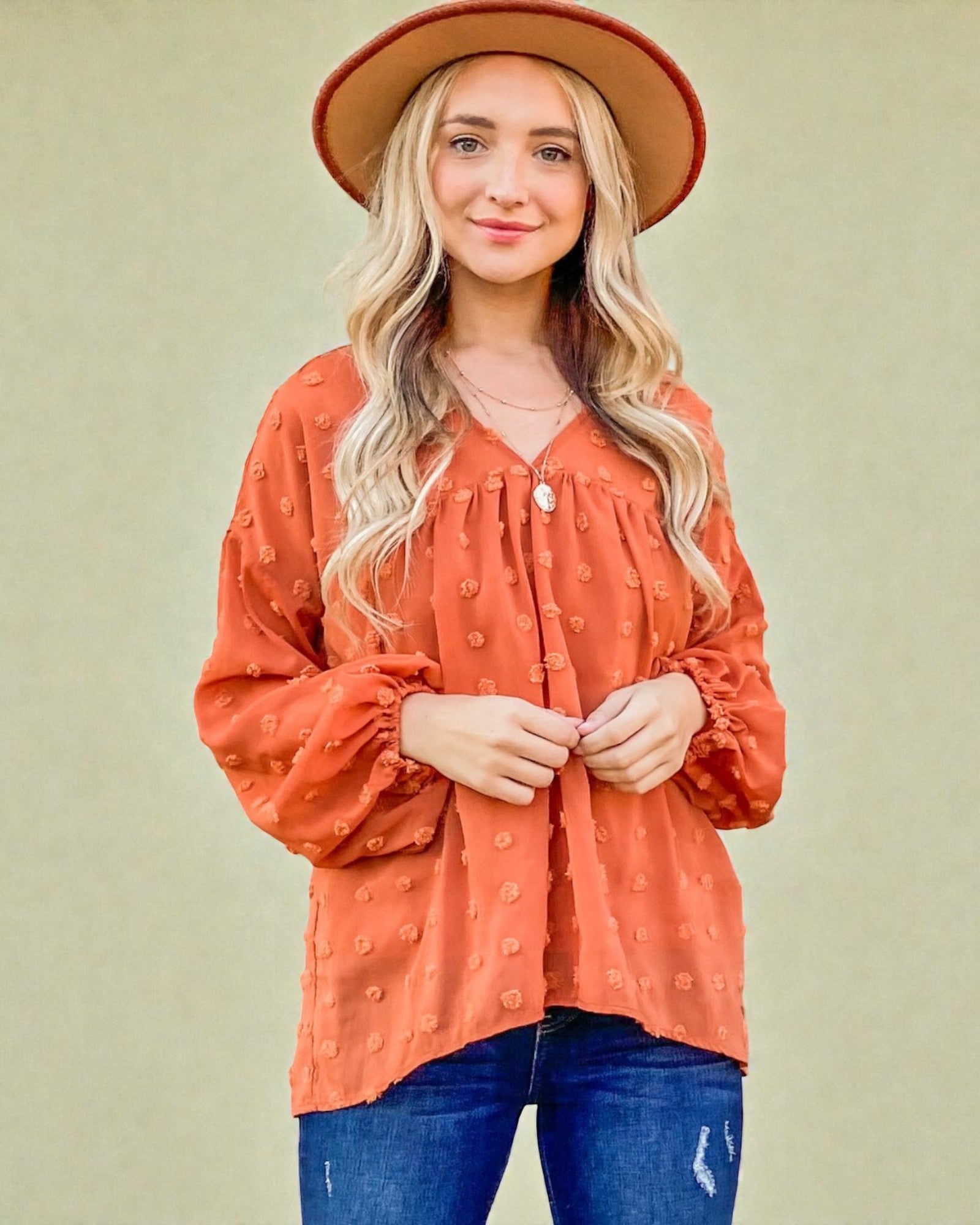 Woman wearing an orange blouse and blue jeans with a beige hat against a beige background