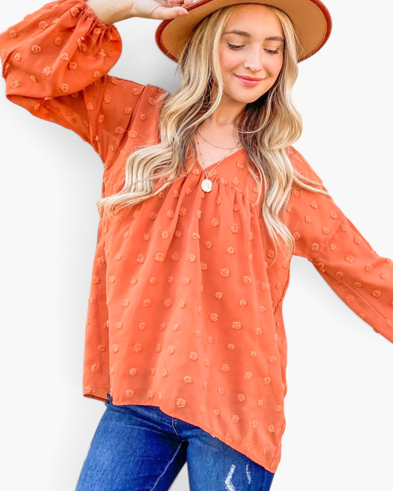 A woman wearing a terracotta orange long-sleeve blouse with a textured, raised dot pattern, paired with blue jeans and a brown wide-brim hat, set against a plain white background.