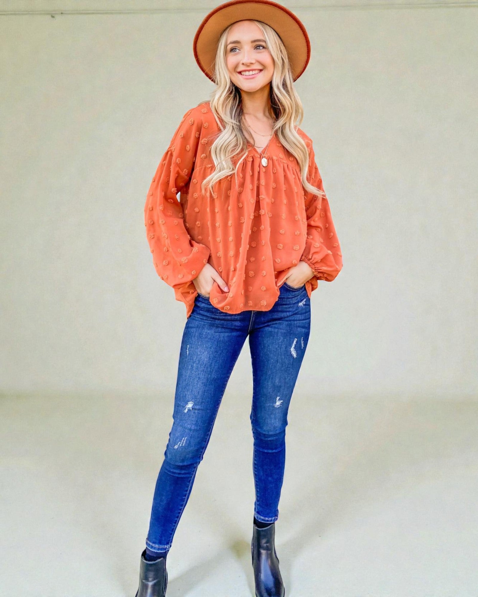 Woman wearing an orange long-sleeve top and blue jeans with a beige hat against a white background