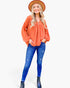 A woman modeling a loose-fitting, long-sleeve orange blouse with a gathered neckline, paired with blue jeans, black ankle boots, and a brown wide-brim hat, against a plain white studio background.