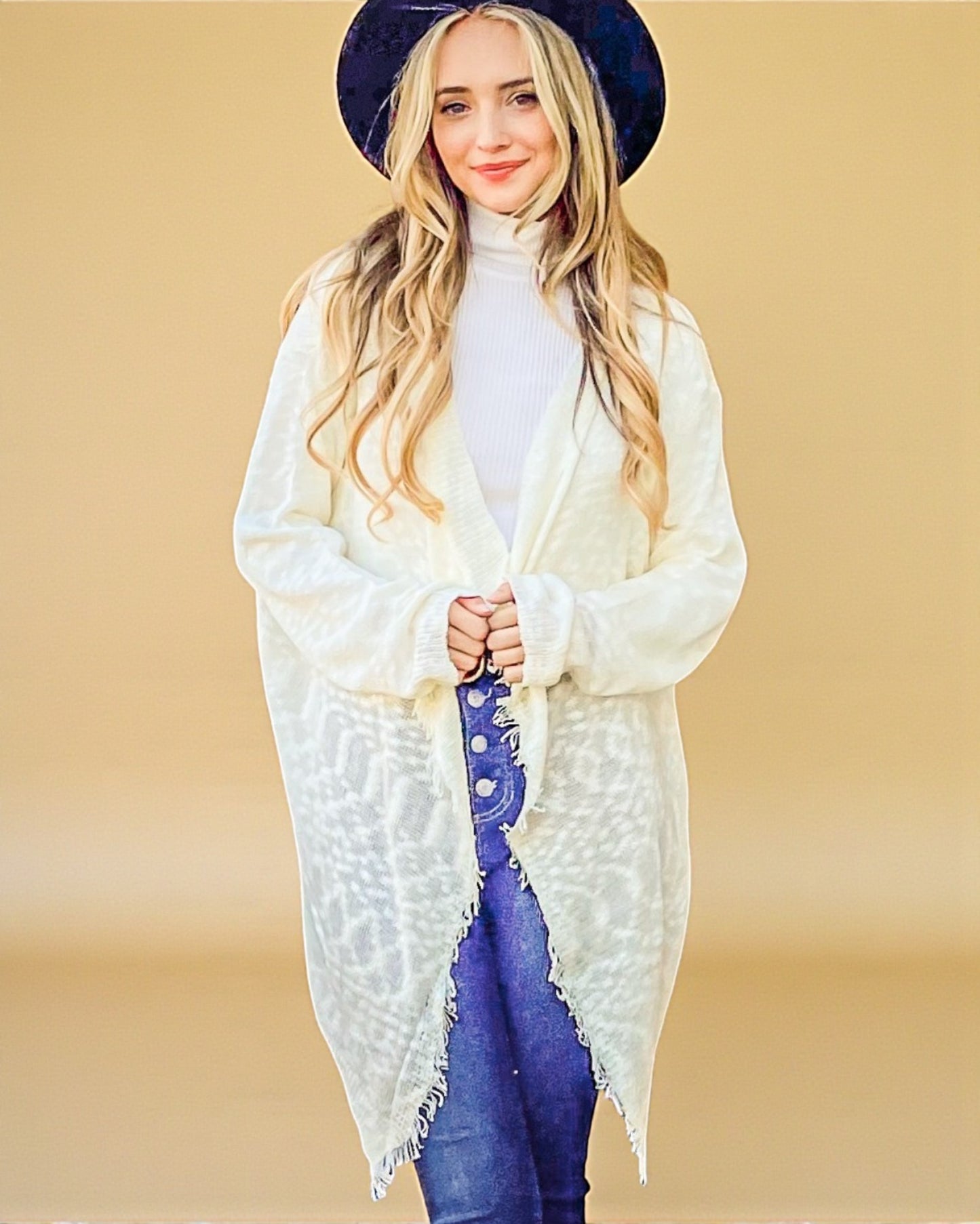 Woman wearing a white cardigan, white turtleneck, blue jeans, and a dark blue hat against a beige background