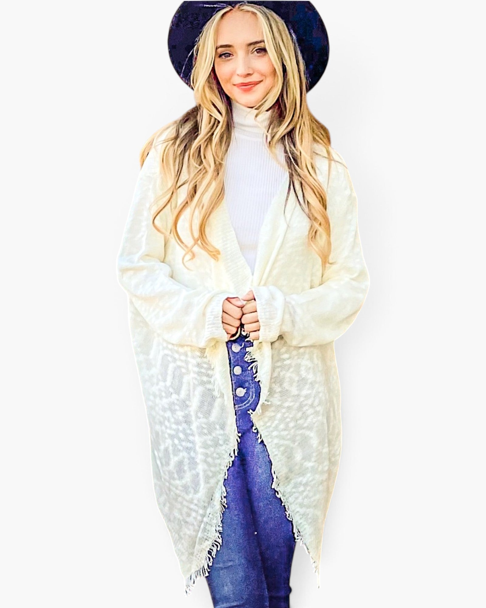 A woman modeling a long cream-colored open-front cardigan with frayed edges, worn over a white turtleneck and blue jeans, while wearing a dark navy wide-brim hat against a plain white background.