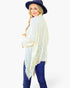 A woman wearing a cream-colored lace-trimmed shawl poncho over a purple top, paired with blue jeans and a navy blue wide-brim hat, photographed against a solid white background.