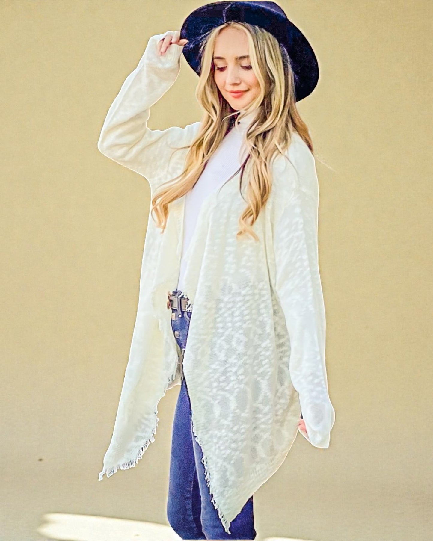 Woman wearing a white cardigan over a white shirt with blue jeans and a dark blue hat against a beige background