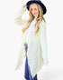A woman modeling a long, open-front cream kimono-style cardigan with a subtle tonal pattern and frayed hems, worn over a white top and blue jeans, while wearing a dark blue wide-brim hat against a plain white background.