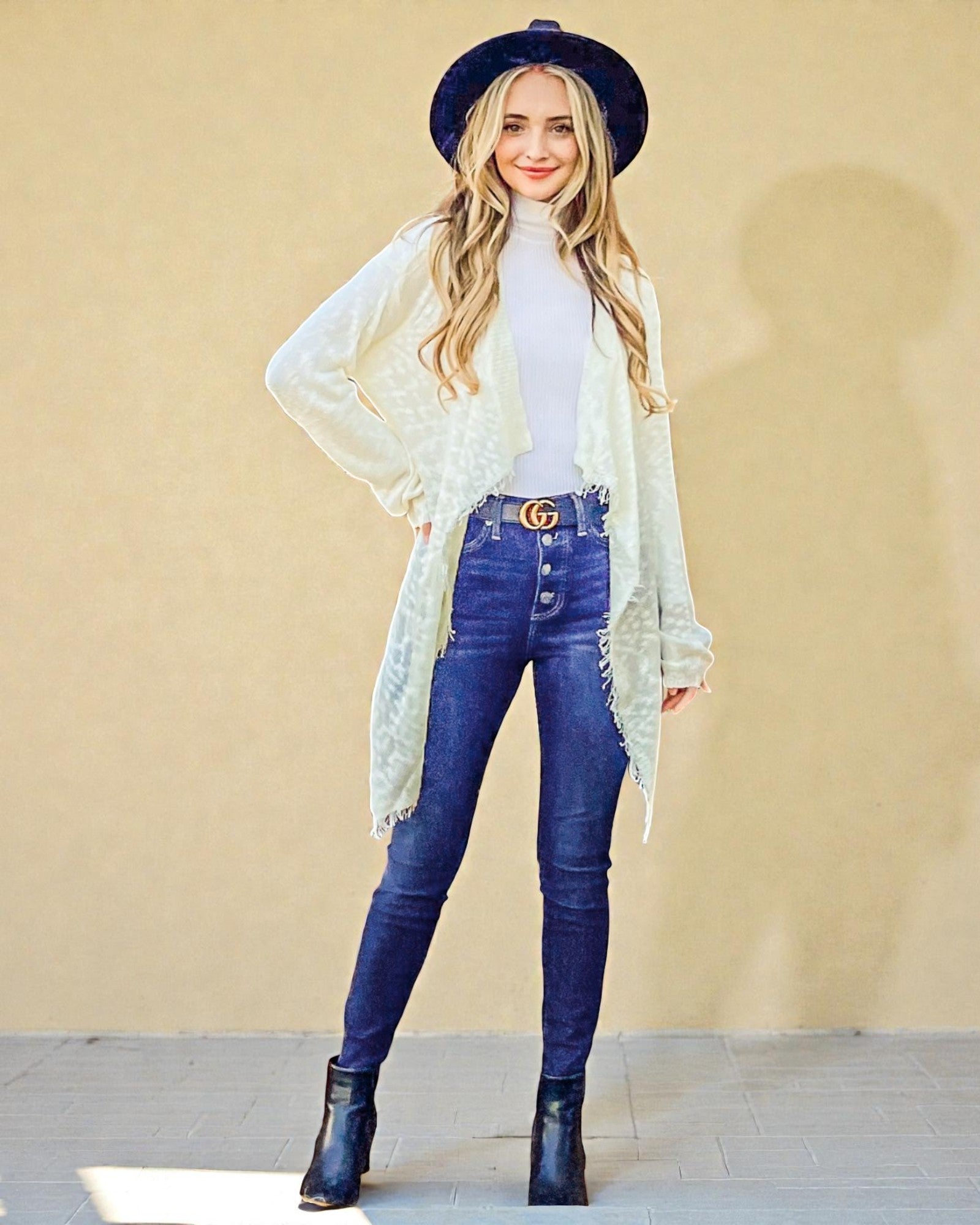 Woman wearing a white lace cardigan, blue jeans, and black boots against a beige wall.
