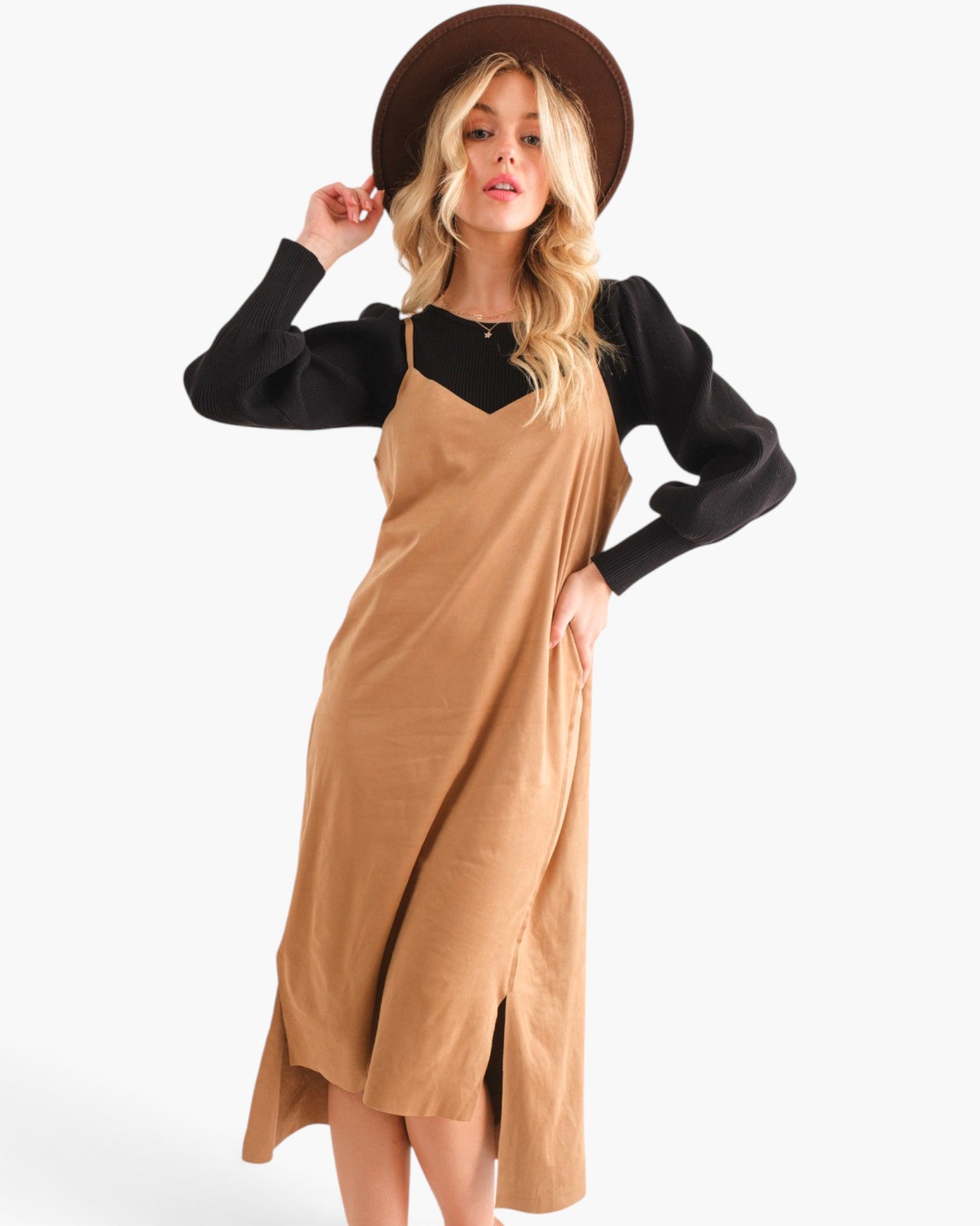 A woman modeling a camel brown linen-look pinafore midi dress over a black long-sleeve top, wearing a brown wide-brim hat, against a plain white studio background.