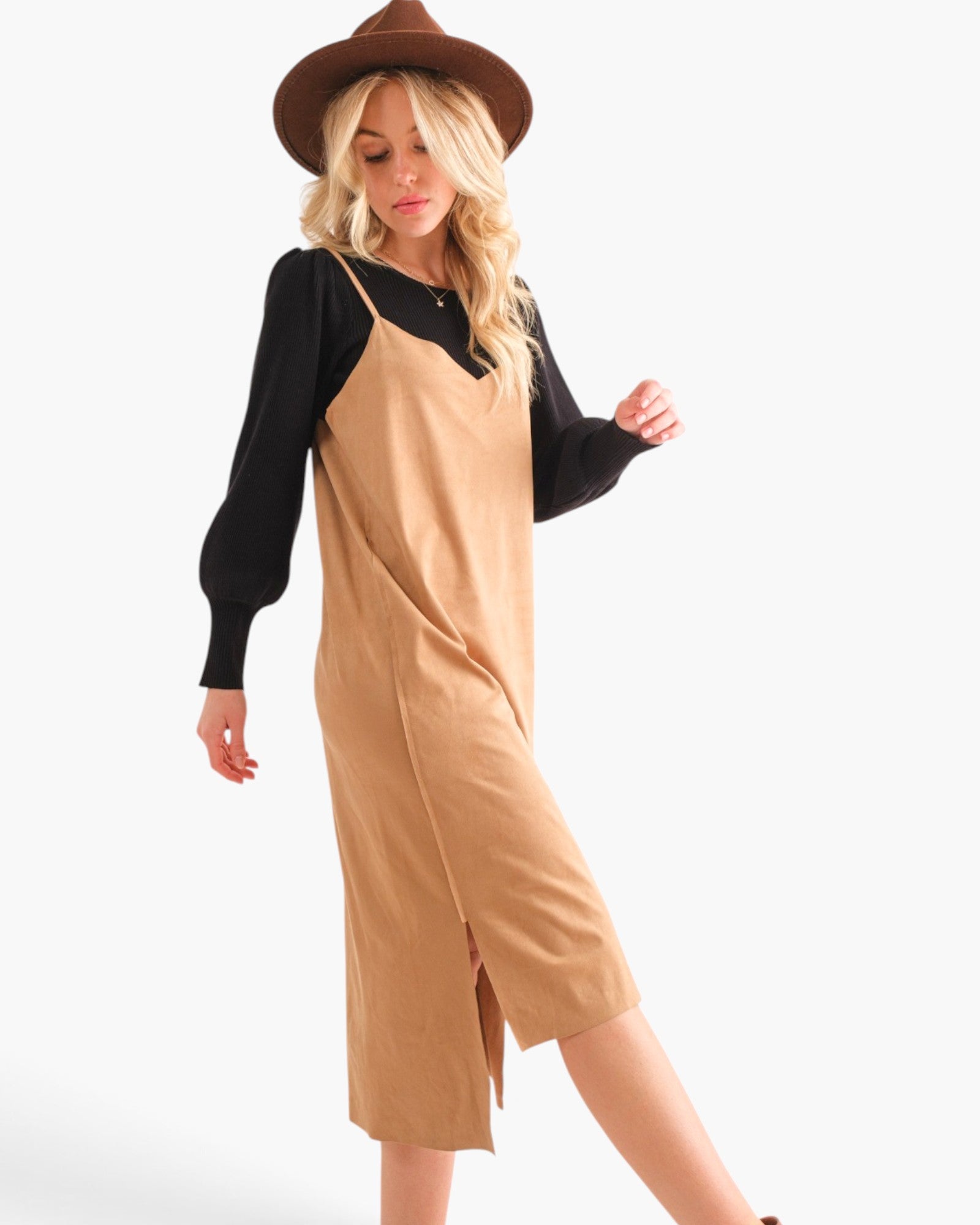 A woman modeling a tan slip-on midi dress layered over a black long-sleeve top, wearing a brown wide-brim hat against a plain white studio background.