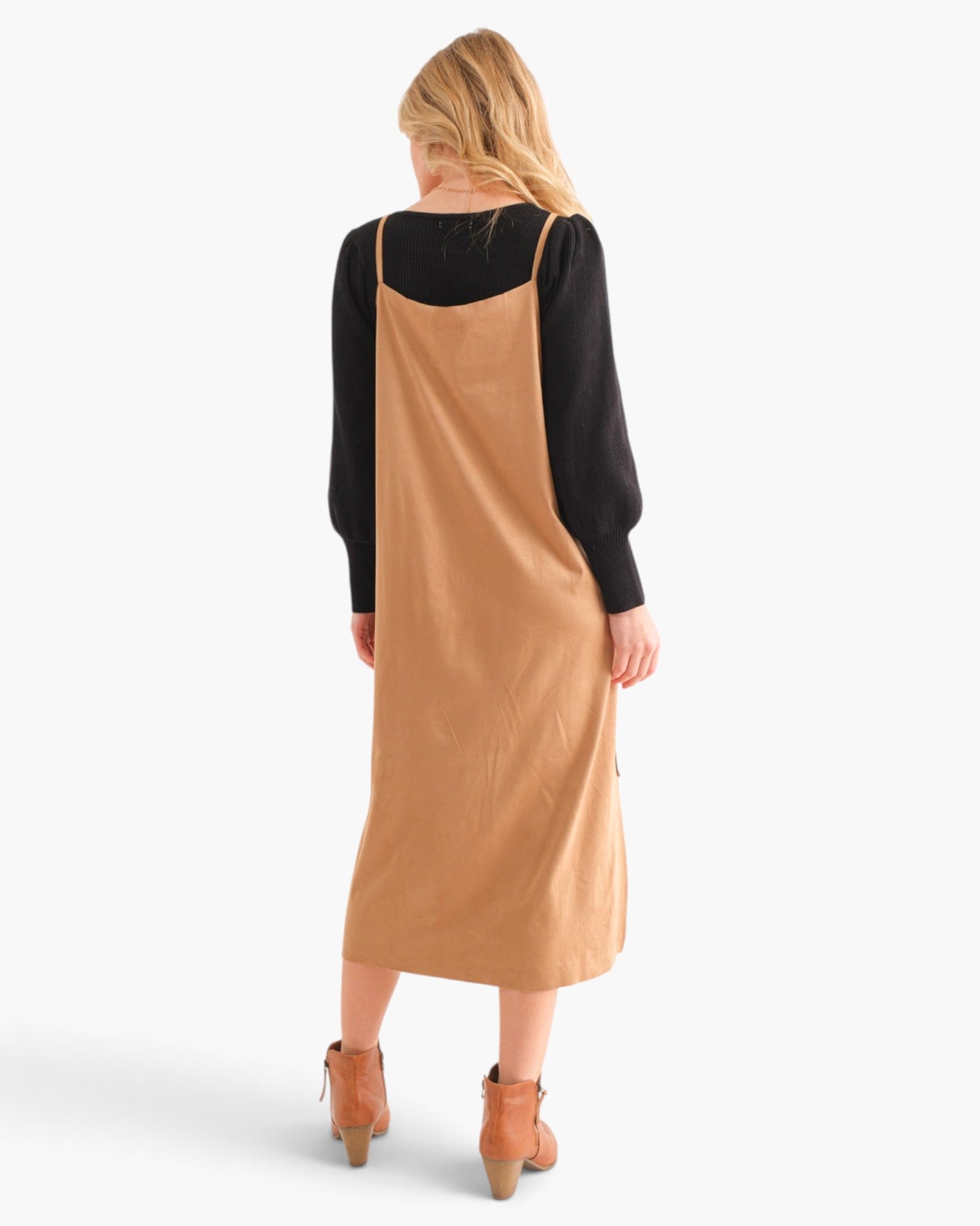 A woman shown from the back wearing a black long-sleeve top under a camel-colored midi slip dress, paired with tan ankle boots, against a light gray studio background.