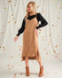 Woman wearing a beige pinafore dress with a black long-sleeve top against a light background with gold dots.