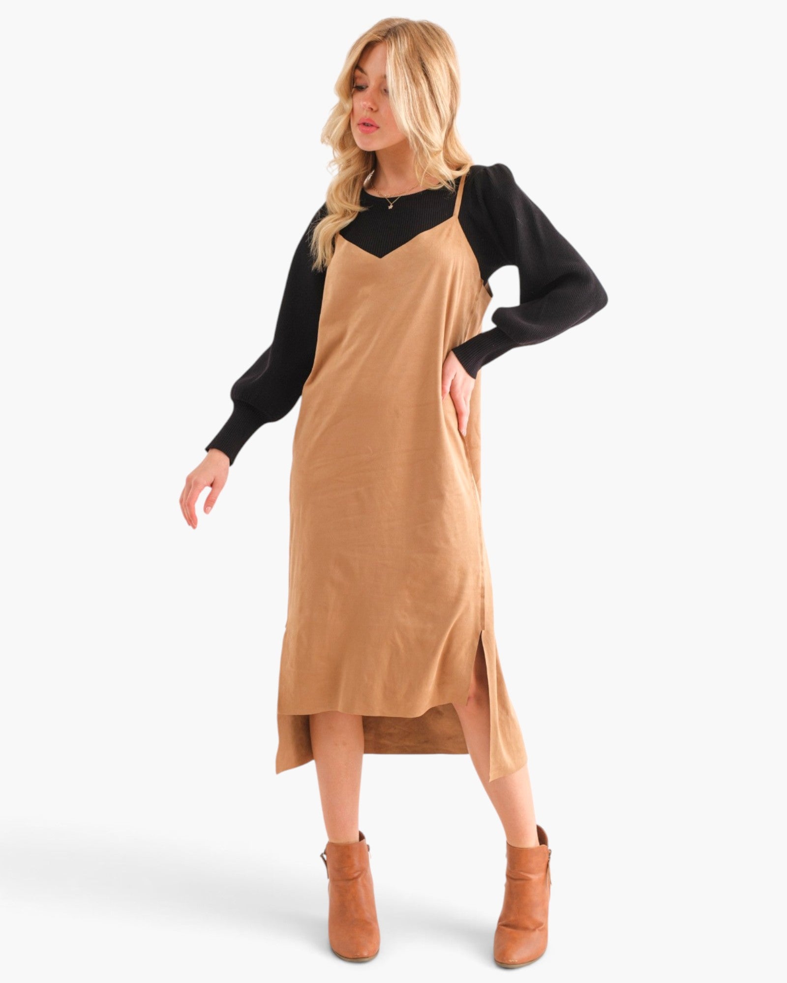 A woman modeling a camel-colored linen slip dress over a black long-sleeve top, paired with tan ankle boots, against a plain white studio background.