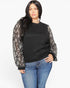 A woman modeling a black crewneck sweatshirt with sheer black-and-white patterned balloon sleeves, paired with blue jeans, against a plain light gray background.