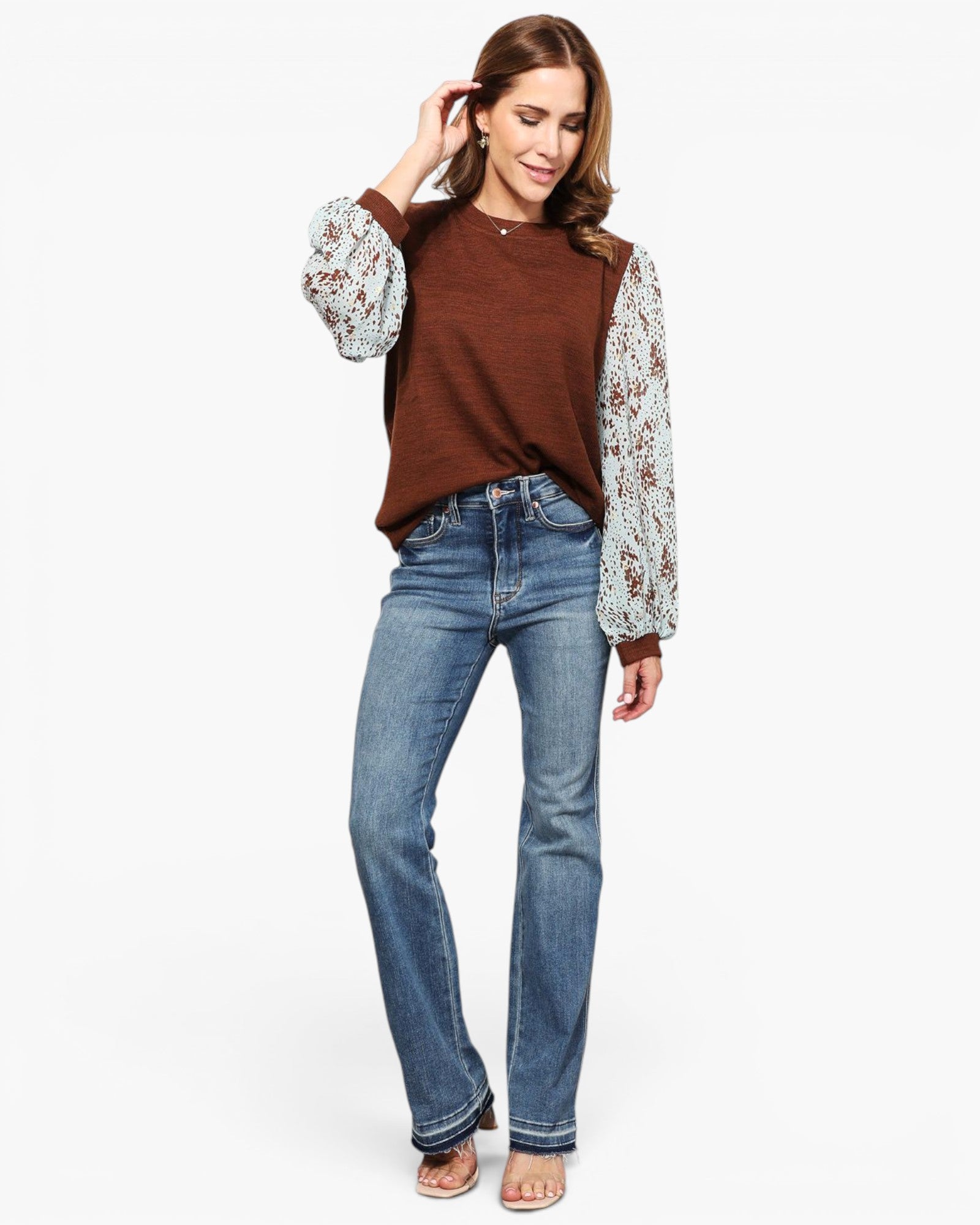 A woman modeling a brown knit sweater vest with long, patterned puff sleeves, paired with blue jeans and flat sandals, against a plain white background.