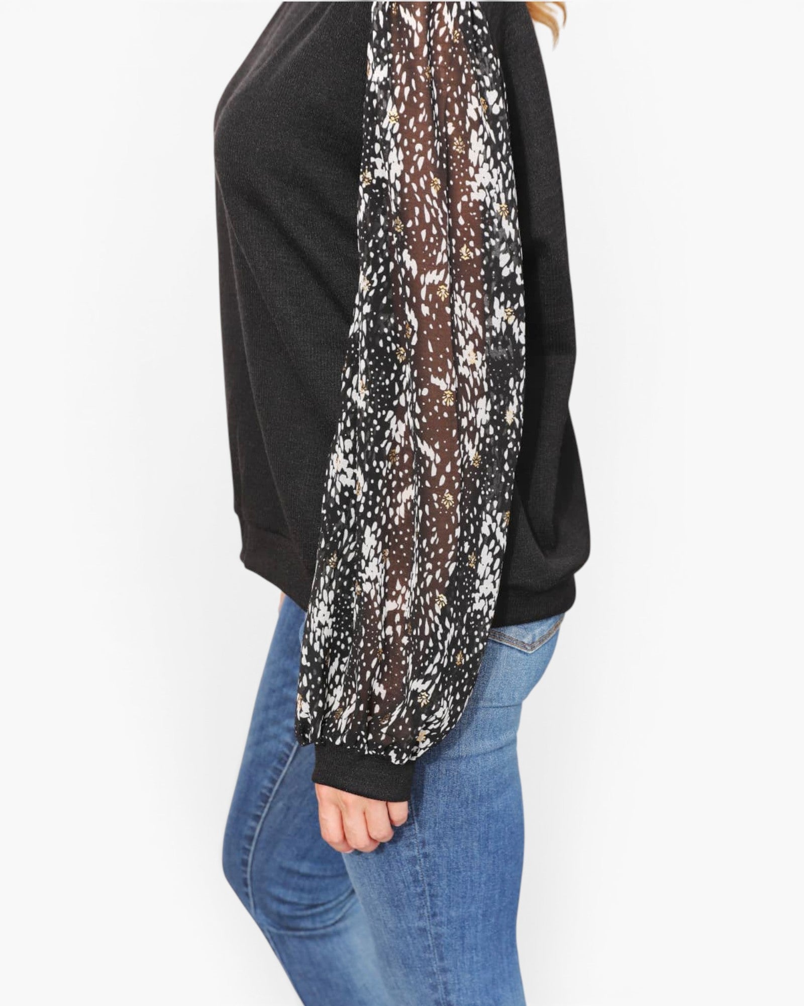 A person wearing a black long-sleeve top with sheer patterned sleeves featuring black, white, and brown spots, paired with blue jeans, shown against a plain white background.
