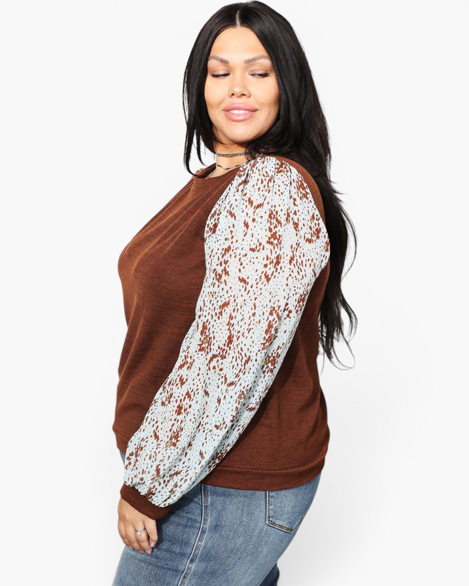 A woman wearing a brown knit top with long white sleeves featuring a brown abstract pattern, paired with blue jeans, photographed against a plain white background.