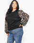 A woman modeling a black long-sleeve top with sheer patterned sleeves, paired with blue jeans, against a plain white background.