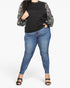 A model wearing a black crewneck top with sheer patterned balloon sleeves, paired with blue skinny jeans and black heeled sandals, shown against a plain light gray studio background.