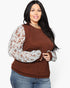 A woman modeling a brown crewneck top with sheer, patterned long sleeves in white and brown, paired with blue jeans against a plain white background.