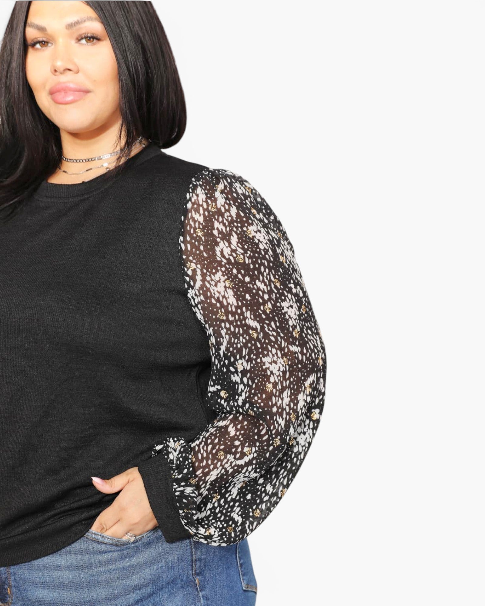 A woman wearing a black long-sleeve top with sheer patterned sleeves, paired with blue jeans, against a plain light gray background.