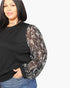 A woman wearing a black long-sleeve top with sheer patterned sleeves, paired with blue jeans, against a plain light gray background.