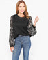 A woman modeling a black crewneck sweater with long, sheer patterned sleeves in black and white, paired with blue jeans against a plain white background.