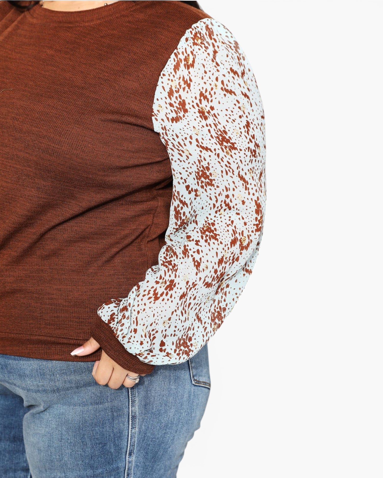 A close-up of a person wearing a brown ribbed knit top with long sheer patterned sleeves in white and rust brown, paired with blue jeans against a plain white background.