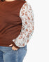 A close-up of a person wearing a brown ribbed knit top with long sheer patterned sleeves in white and rust brown, paired with blue jeans against a plain white background.