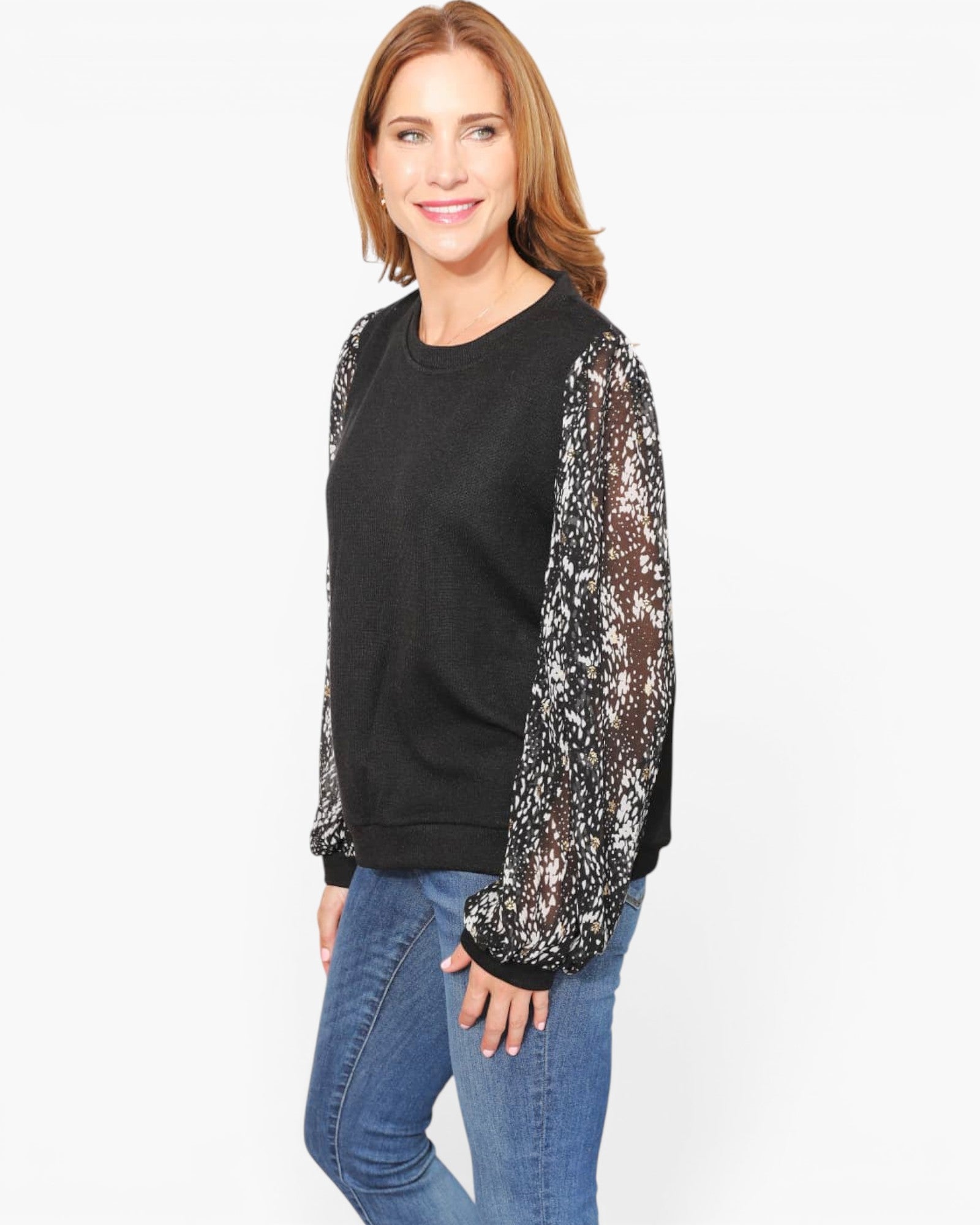 A woman modeling a black crewneck sweatshirt with sheer black-and-white patterned lace sleeves, paired with blue jeans, against a plain white studio background.
