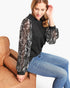 A woman wearing a black crewneck sweatshirt with long sheer black sleeves covered in white sequins, paired with blue jeans, sitting on a brown leather couch against a white background.