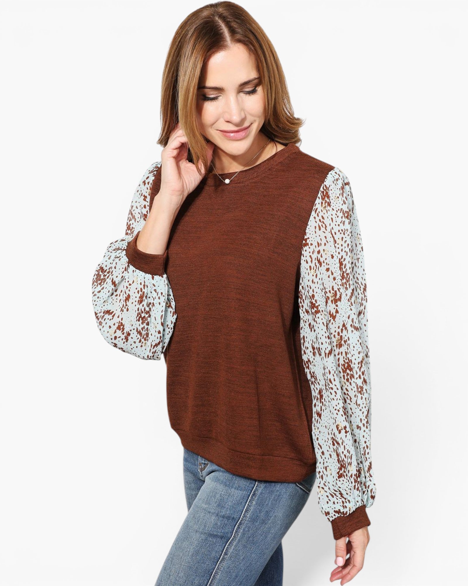 A woman wearing a brown crewneck sweater with long sleeves featuring a white and brown floral lace overlay, paired with blue jeans, against a plain white background.
