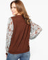 A woman shown from the back wearing a brown sleeveless vest over a white long-sleeve blouse with a brown floral print, paired with blue jeans, against a plain white background.