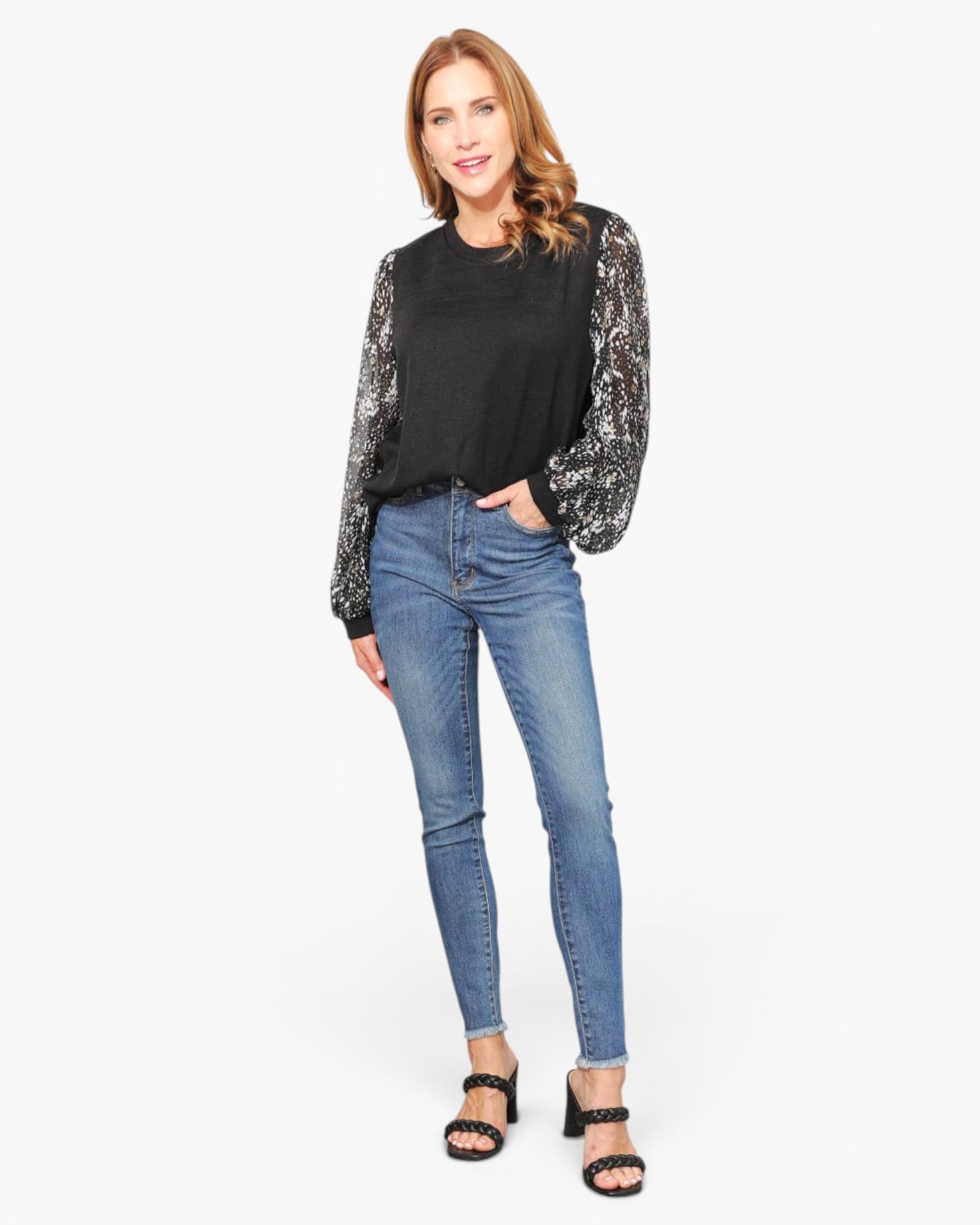 A woman modeling a black long-sleeve top with sheer patterned sleeves, paired with blue skinny jeans and black heeled sandals, against a plain white studio background.
