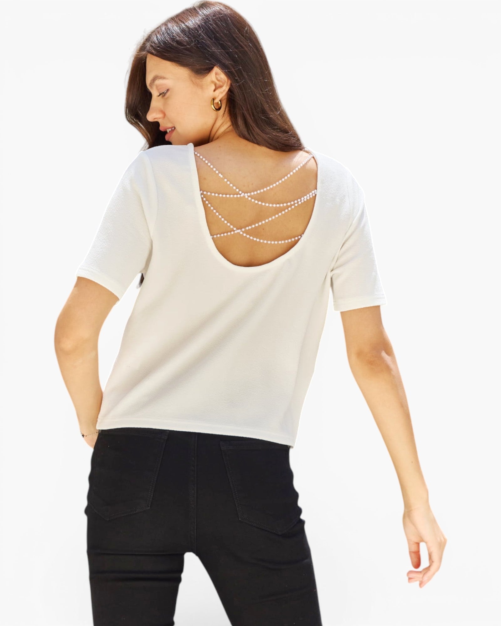 A woman shown from the back wearing a cream-colored short-sleeve top with a decorative open back featuring crisscrossing pearl chains, paired with black pants, against a plain white background.