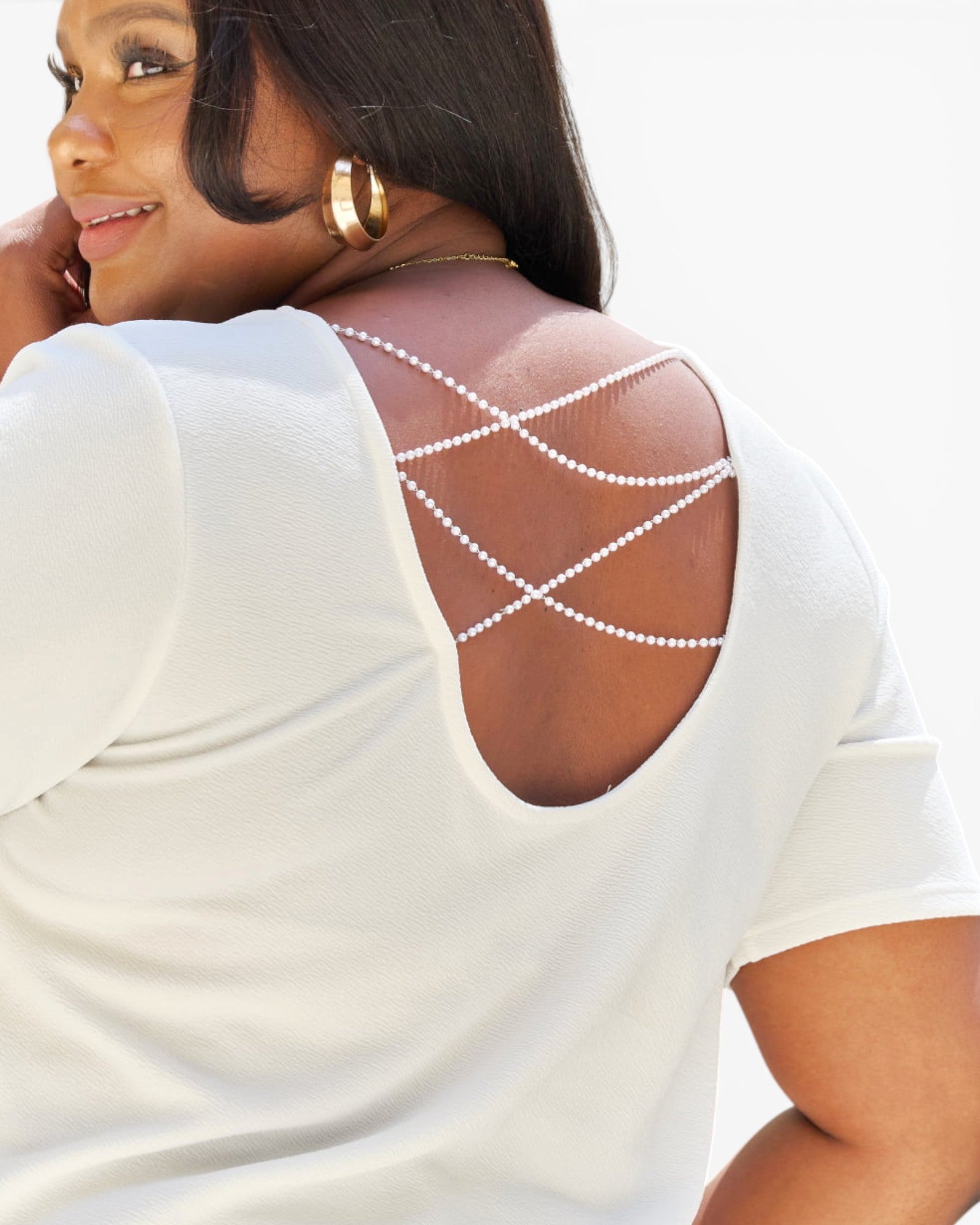 A close-up back view of a woman wearing a white short-sleeve top with decorative beaded crisscross straps across the open back, set against a bright white background.