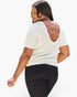 A woman wearing a white short-sleeve top with an open back and crisscross chain detail, paired with black pants, shown from a three-quarters back view against a light gray studio background.
