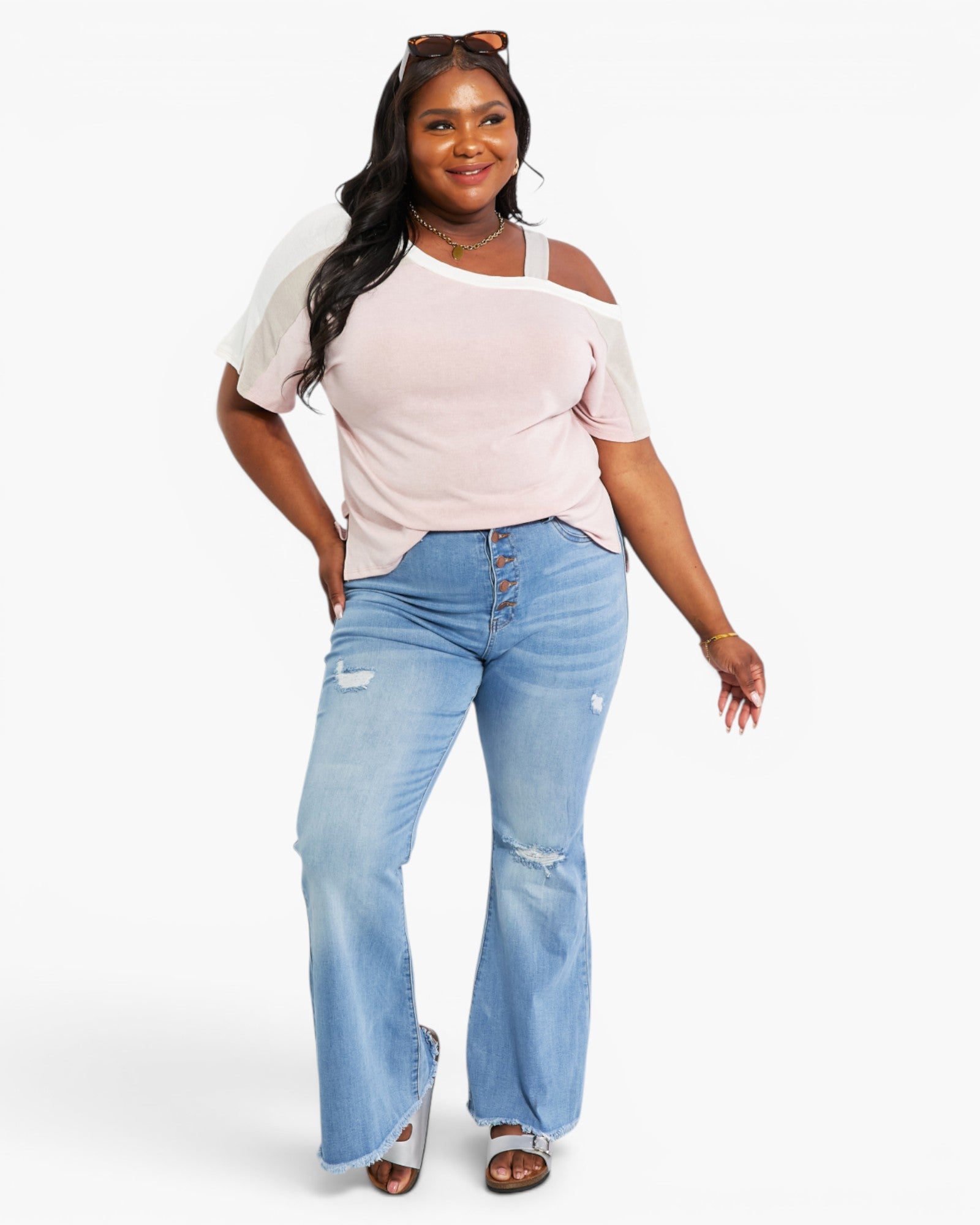 A woman modeling a light pink cold-shoulder t-shirt with sheer white sleeves and light-wash blue distressed flare jeans, standing against a plain white studio background.