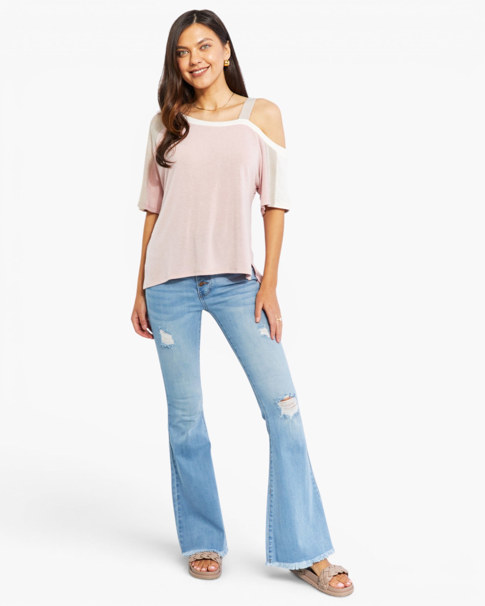 A woman modeling a pale pink cold-shoulder top with white sleeves and light-wash blue distressed flare jeans, standing against a plain white studio background.