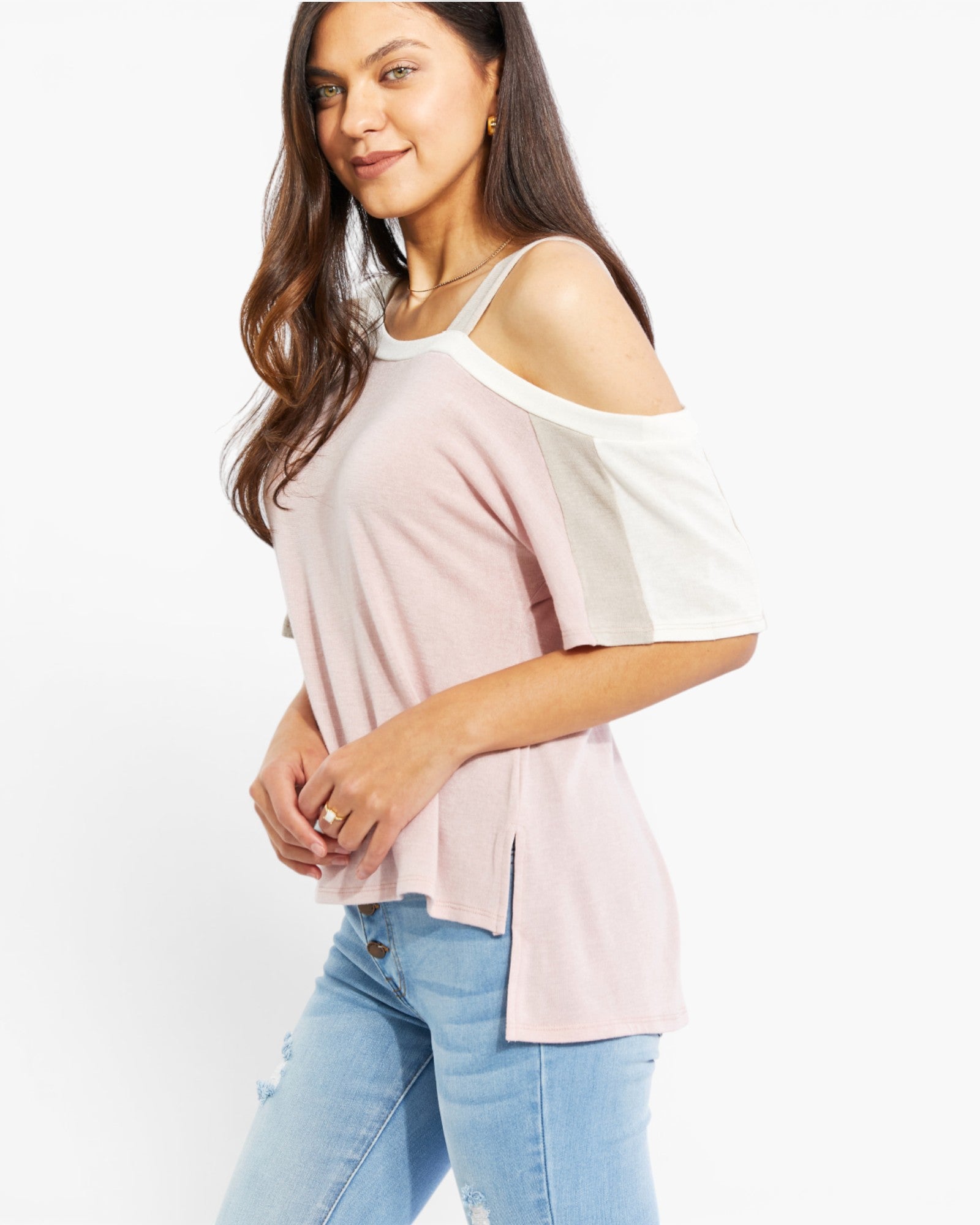 A woman wearing a light pink cold-shoulder top with white shoulder panels and light blue jeans, photographed against a plain white studio background.