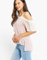 A woman wearing a light pink cold-shoulder top with white shoulder panels and light blue jeans, photographed against a plain white studio background.