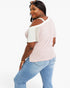 A woman modeling a light pink cold-shoulder t-shirt with contrasting white shoulder panels and light blue jeans, photographed against a solid white studio background.