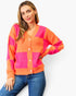 A woman wearing a bright orange and hot pink checkered knit cardigan sweater with blue jeans, posing against a light gray studio background.